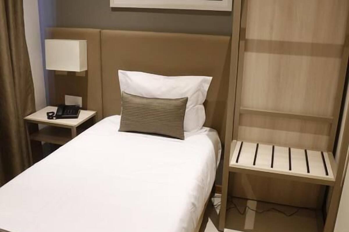 A hotel room with a bed, bedside table, lamp, and a wooden shelf unit.