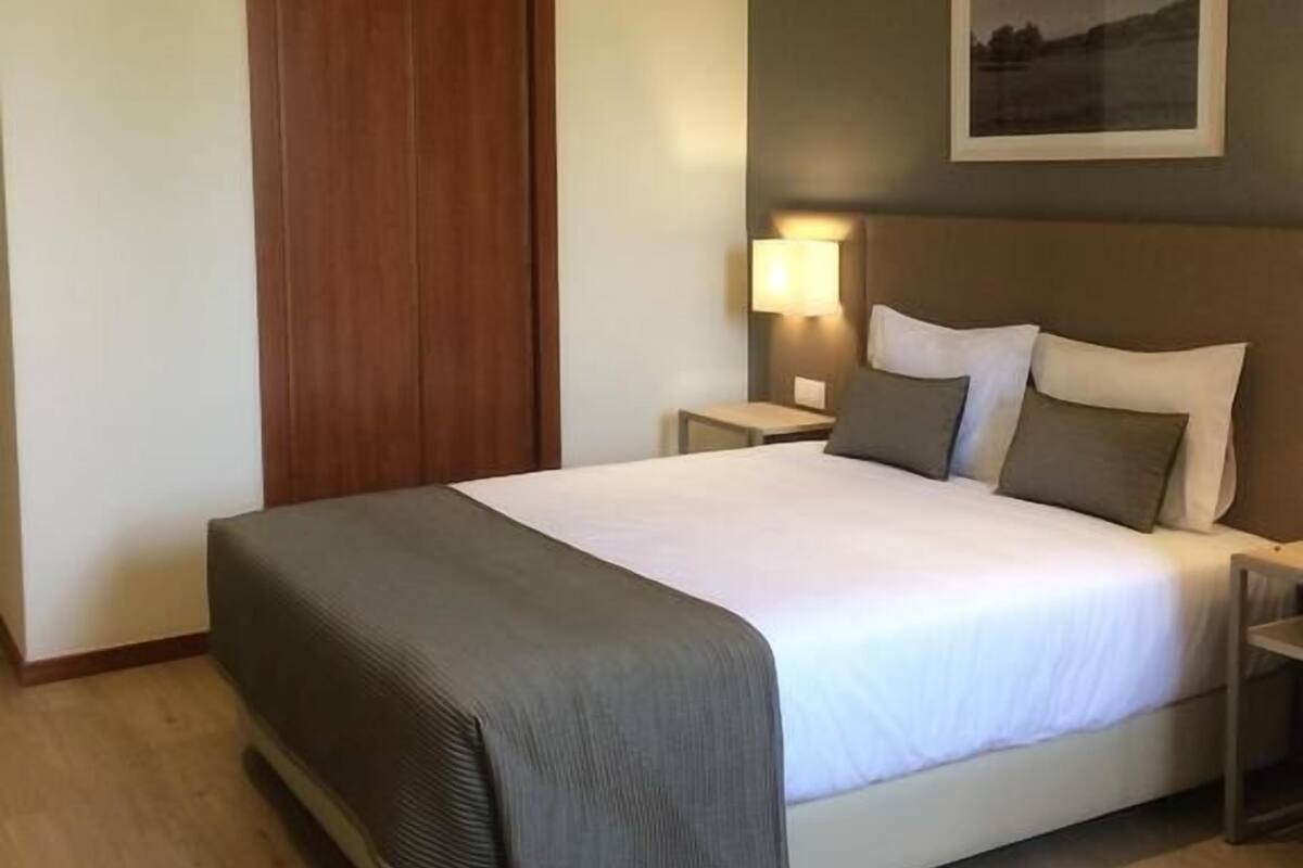 A hotel room with a large bed, a wooden door, a nightstand with a lamp, and a framed picture on the wall.