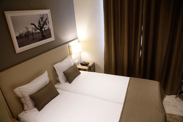 A hotel room with a large bed, a bedside table with a lamp, a framed picture on the wall, and a window with curtains.