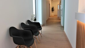 Un couloir avec des chaises noires alignées contre un mur blanc, menant à une pièce où se trouvent une plante et une porte.
