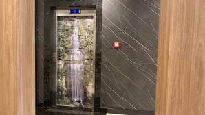 Un ascensor con pantalla digital, botón de emergencia rojo y vistas a una cascada.