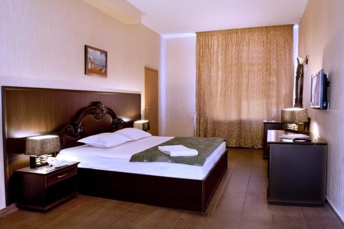 A hotel room with a large bed, a wooden headboard, a bedside table with a lamp, a small desk with a lamp, and a television on the wall.