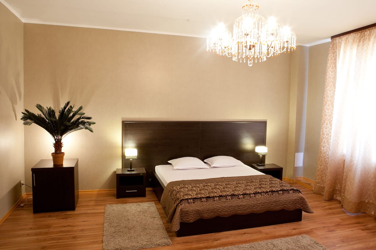 A bedroom with a large bed, two bedside tables with lamps, a chandelier, a potted plant, and a window with curtains.
