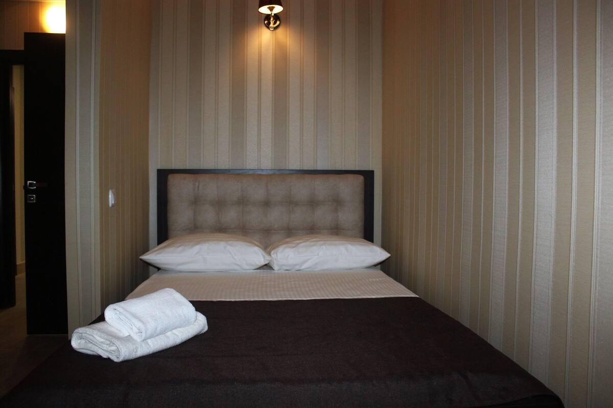 A neatly made bed with a dark headboard and white pillows, a folded white towel on the bed, and a wall-mounted light fixture above.
