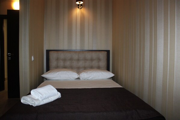 A neatly made bed with a dark headboard and white pillows, a folded white towel on the bed, and a wall-mounted light fixture above. - Hotel Verhovina (Petropavlivs'ka Borshchahivka)