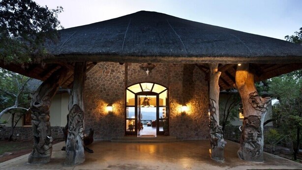 A stone pavilion with a thatched roof, supported by carved tree trunks, and a view into a warmly lit interior. - Izingwe Lodge (Vaalwater)