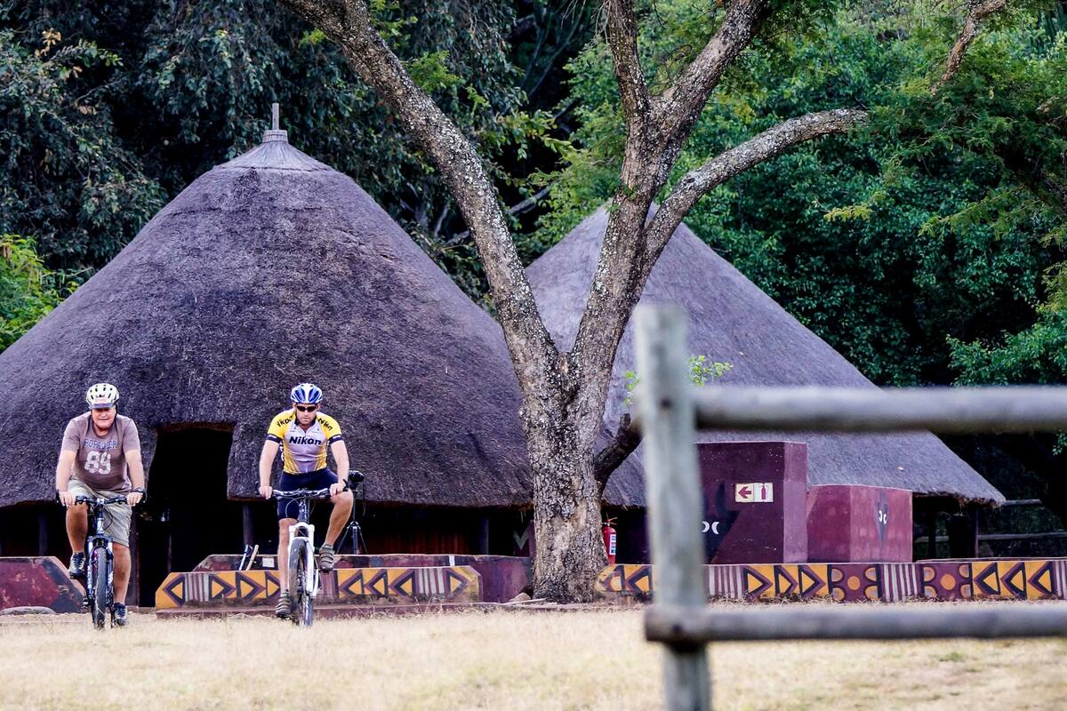 Two cyclists in front of thatched-roof huts with a tree and a metal fence in the foreground.