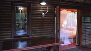 Ein rustikales Holzhütten-Interieur mit einem Bett unter einem Moskitonetz, einem Holztisch und einer Deckenleuchte.