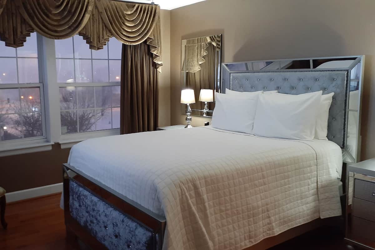 Une chambre d’hôtel avec un grand lit, une table de chevet avec une lampe, une chaise et une fenêtre avec des rideaux.