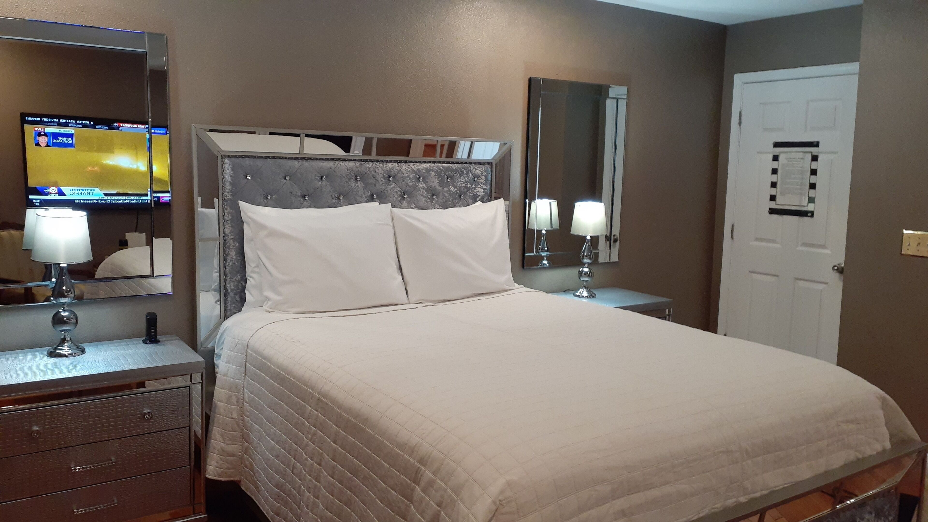 A neatly made bed with a tufted headboard, two bedside lamps, a dresser with drawers, a large mirror, and a television on the wall.