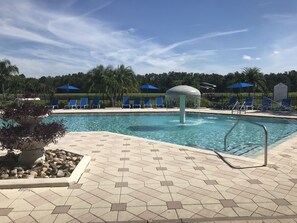 Piscina con sillas y sombrillas azules, rodeada por un suelo de baldosas y una fuente.