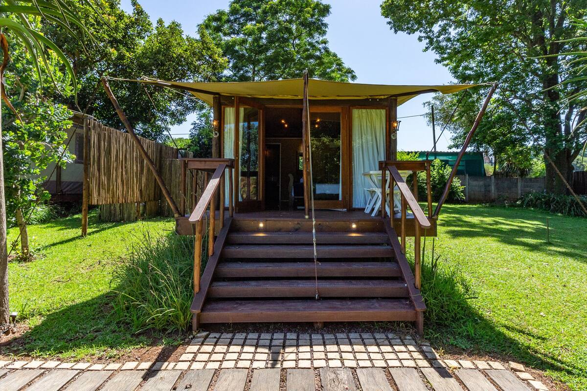 A wooden staircase leading to a covered entrance of a building, surrounded by lush greenery and a brick pathway.