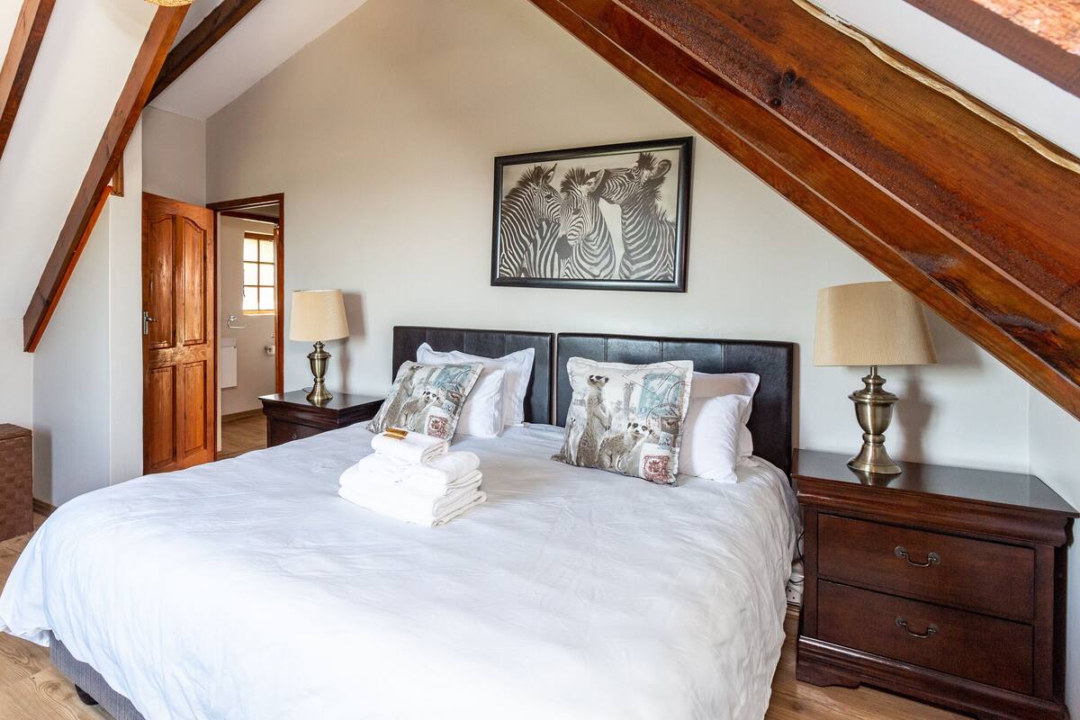 A bedroom with a large bed, wooden headboard, and a wooden ceiling. There is a door leading to a bathroom, a nightstand with a lamp, and a dresser.