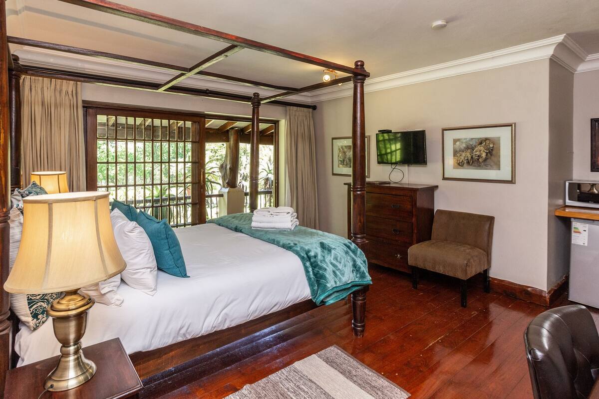 A four-poster bed with a wooden headboard, a bedside table with a lamp, a chair, a desk with a microwave, and a television on the wall.