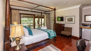 A four-poster bed with a wooden headboard, a bedside table with a lamp, a chair, a desk with a microwave, and a television on the wall. - Kangelani Lodge (Assagay)