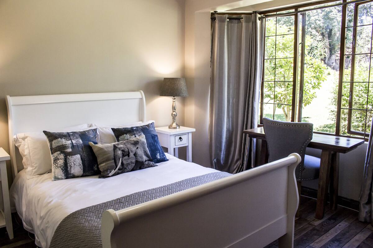 A neatly made bed with a white headboard, a bedside table with a lamp, and a view of greenery through large windows.