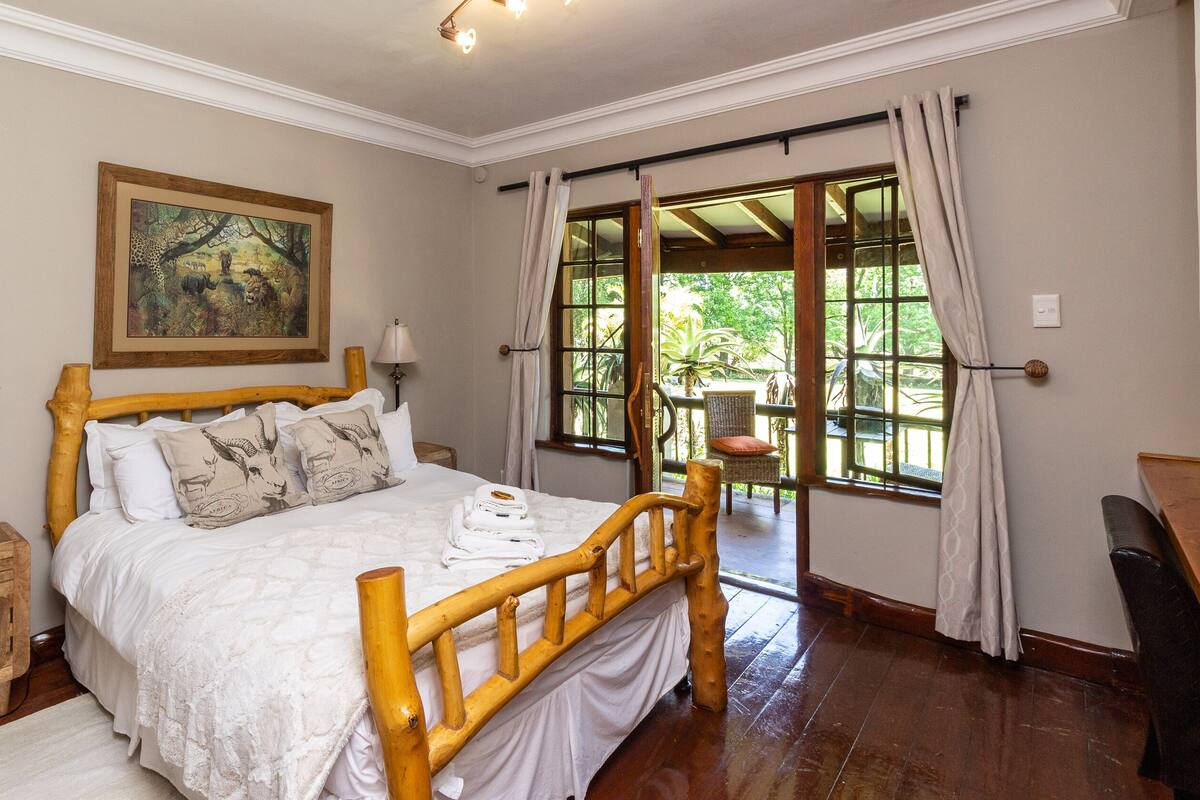A bedroom with a wooden bed, a painting on the wall, a window with curtains, and a view of greenery outside.