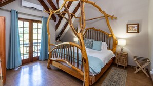 A rustic bedroom with a four-poster bed, wooden headboard, and a view of the outdoors through a large window. - Kangelani Lodge (Assagay)
