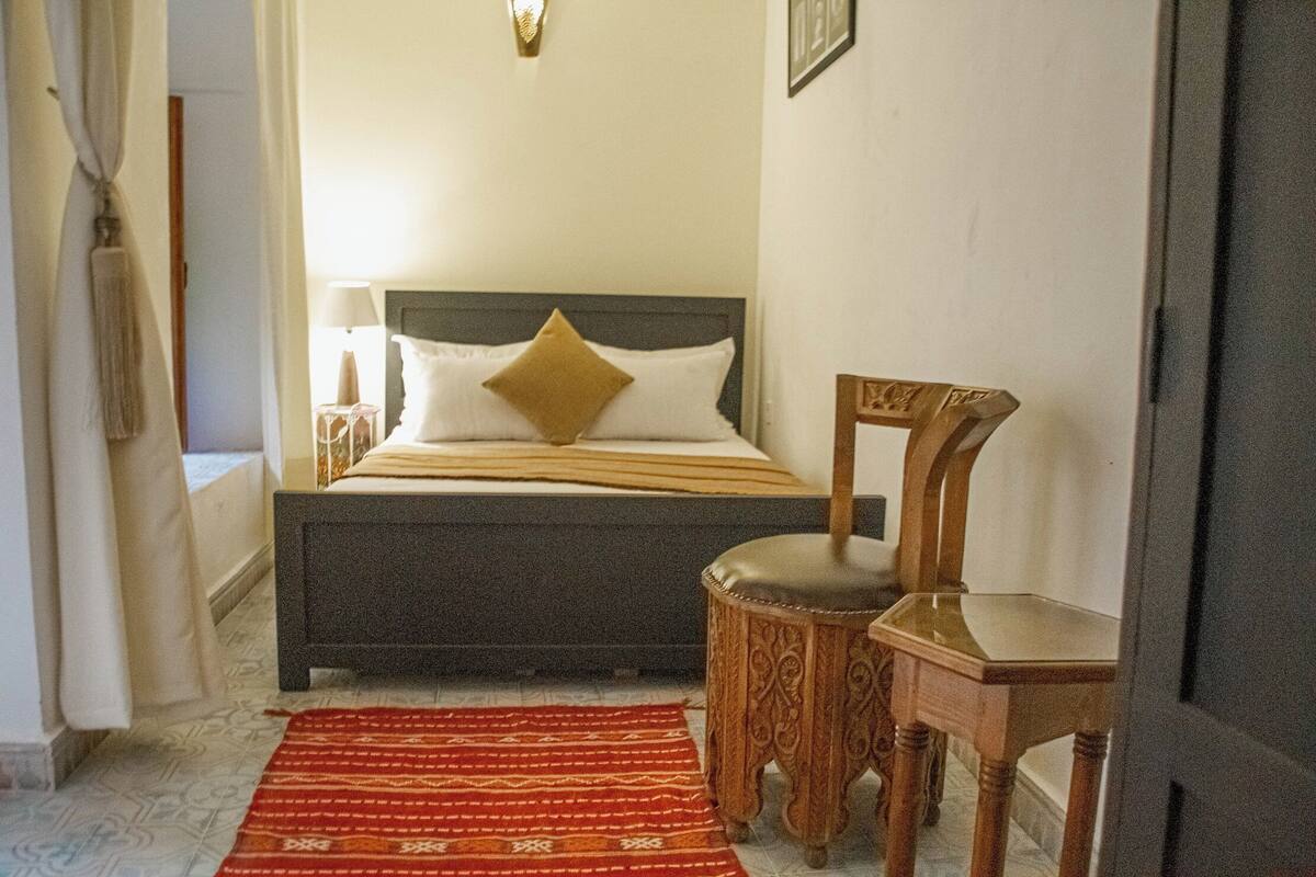 Un dormitorio con cama, una silla, un taburete pequeño y un tapete estampado.