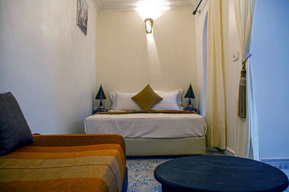 Una habitación de hotel con una cama, dos mesitas de noche, una mesita redonda pequeña y un suelo estampado.