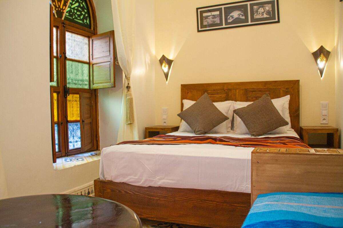 Una cama bien hecha con tablero de madera, dos almohadas y una manta a rayas. Hay dos mesitas de noche con lámparas, una ventana con cortinas y un cuadro colgado en la pared.