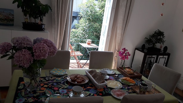 Un comedor con una mesa dispuesta para el desayuno, que incluye pan, bollería y fruta. Hay sillas alrededor de la mesa y se ve una planta en maceta al fondo.