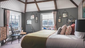 A bedroom with a large bed, a view of the city through a window, and a chair by the window. - Chainlocker (Falmouth)