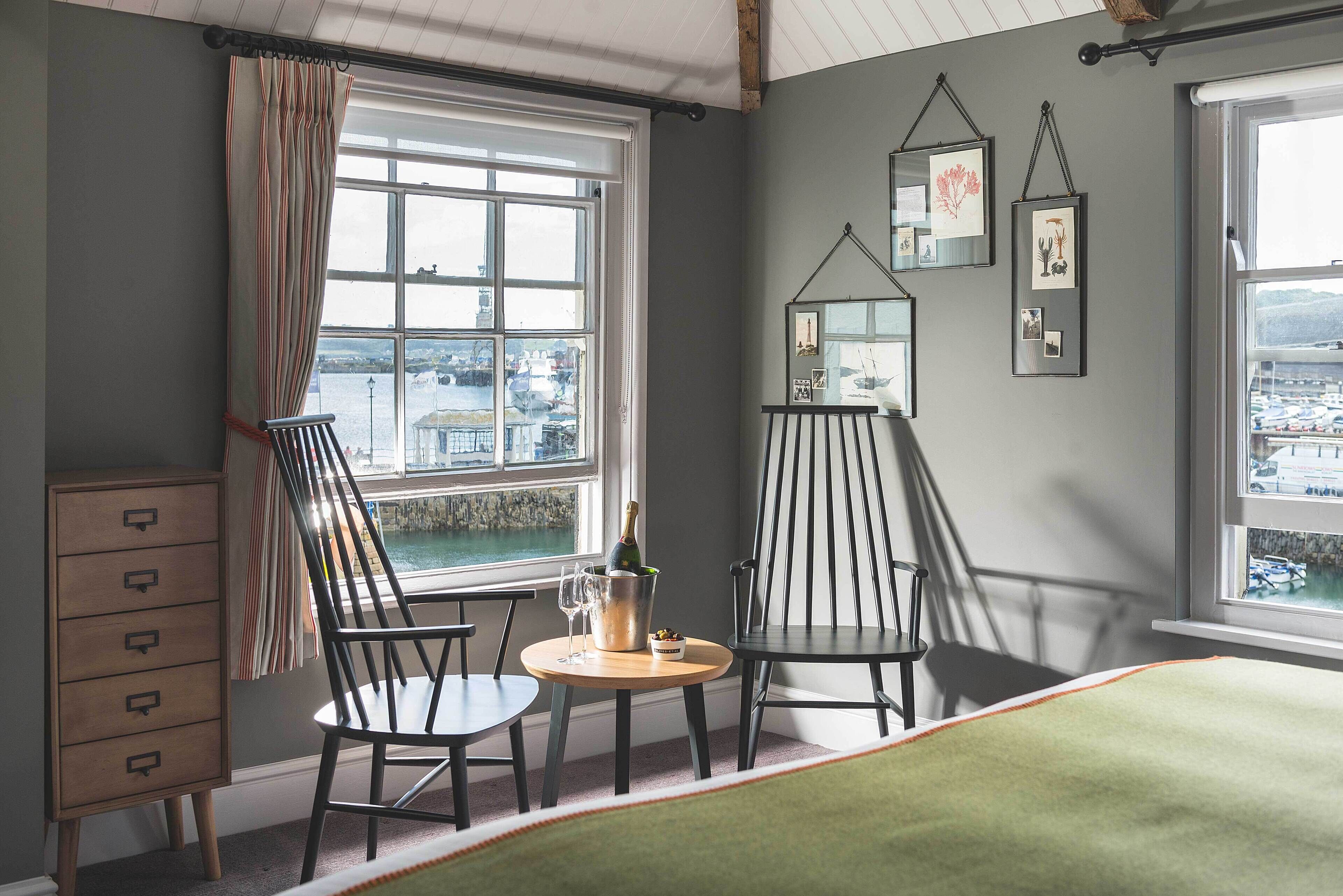 A room with a view of a harbor, two chairs, a small table with a bottle and glasses, and a dresser.