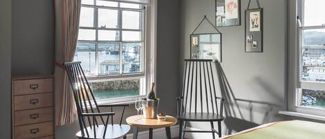 A room with a view of a harbor, two chairs, a small table with a bottle and glasses, and a dresser.