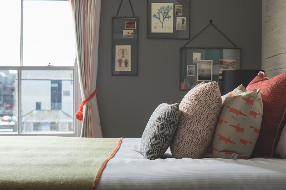A neatly made bed with patterned pillows, a green throw blanket, and a view of a cityscape through a window.