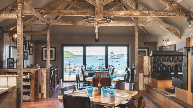 A restaurant with exposed wooden beams, a view of the sea, and a bar area with stools. - Chainlocker (Falmouth)
