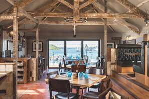 A restaurant with exposed wooden beams, a view of the sea, and a bar area with stools. - Chainlocker (Falmouth)
