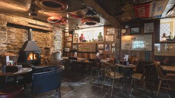 Un pub acogedor con pared de piedra, techo de madera y chimenea. Hay varias mesas y sillas de madera, y una zona de bar con mostrador y taburetes.