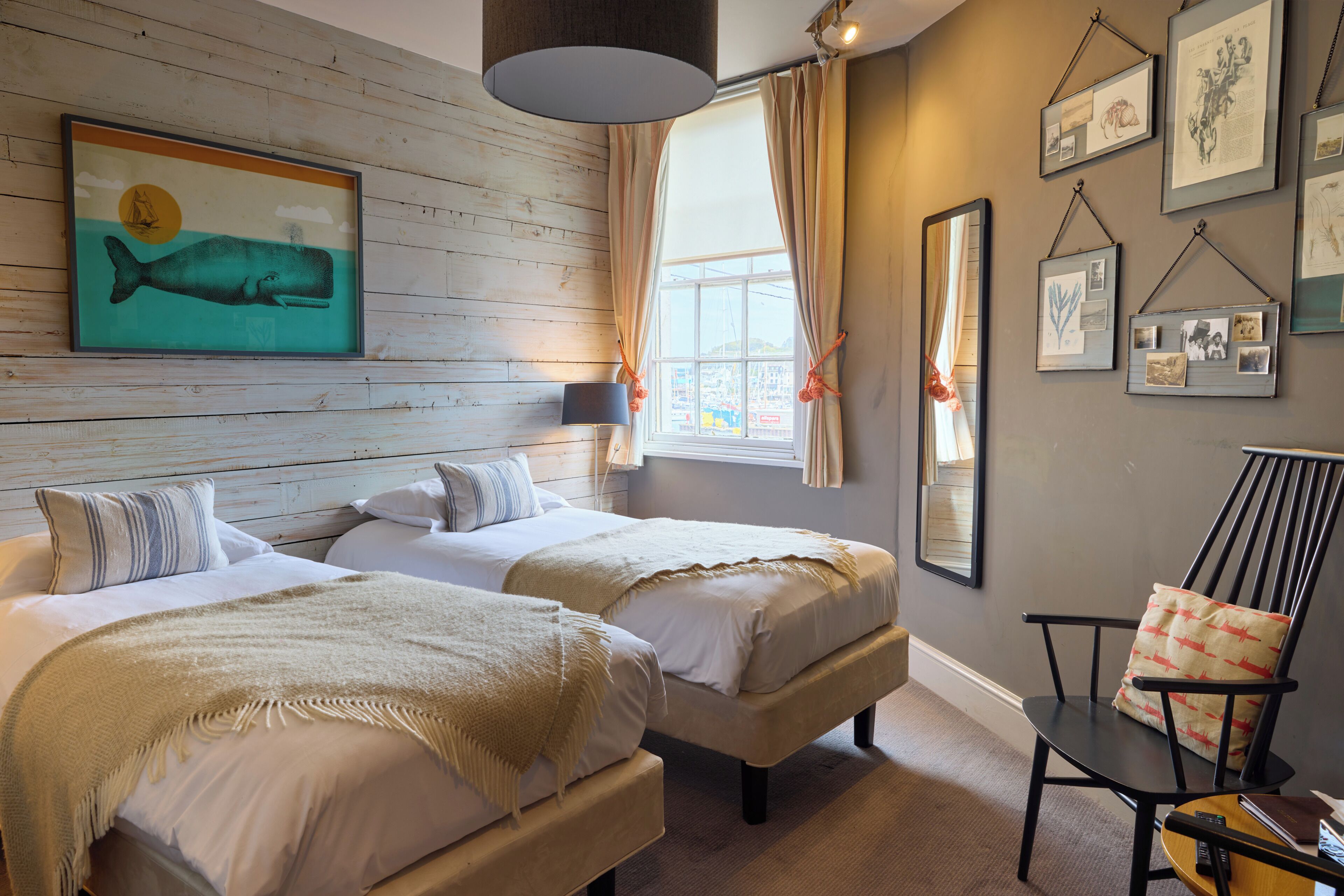 A hotel room with two beds, a large window with curtains, a wall-mounted lamp, and various framed pictures.