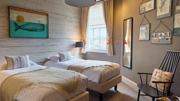 A hotel room with two beds, a large window with curtains, a wall-mounted lamp, and various framed pictures.