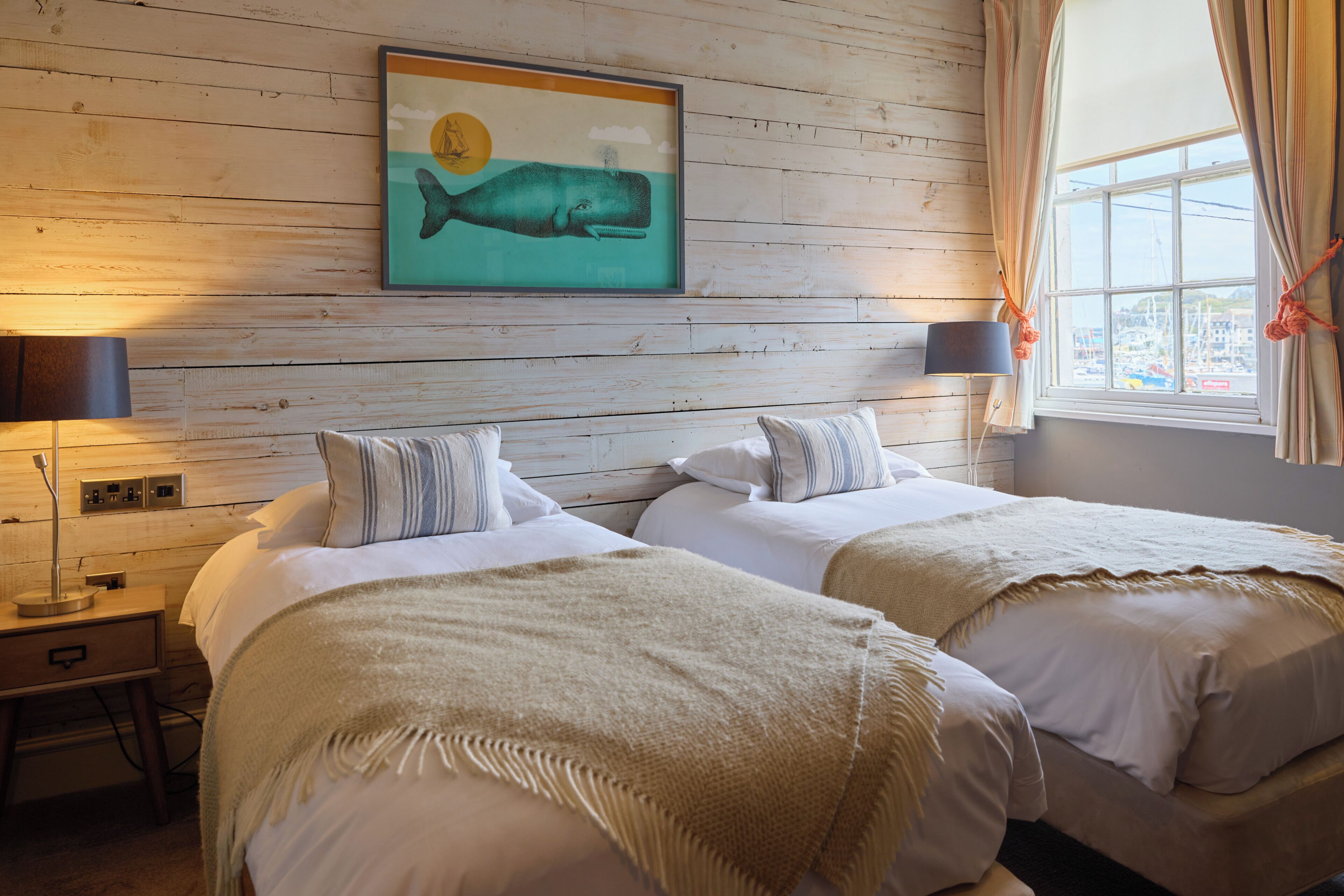 A room with two beds, a nightstand, a lamp, a painting of a whale, and a window with curtains.
