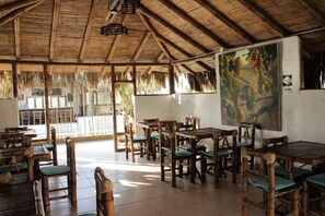 A restaurant with wooden tables and chairs, a thatched roof, and a painting of grapes on the wall. - Puerto Antiguo (Los Organos)