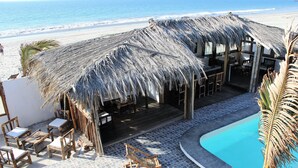 Un complexe hôtelier en bord de mer doté d’un espace extérieur avec des meubles en bois et une pergola en chaume, ainsi que d’une piscine.