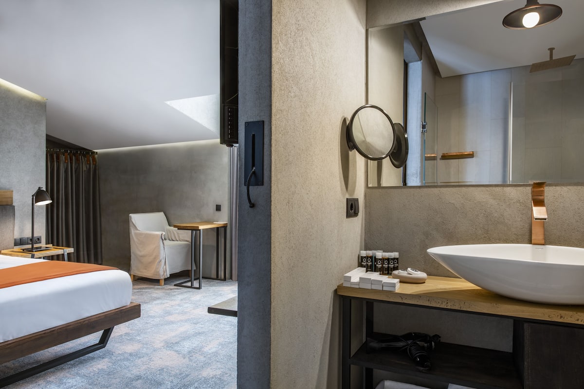 A modern hotel room with a large bed, a small desk, a chair, a large mirror, and a bathroom with a large sink and shower.