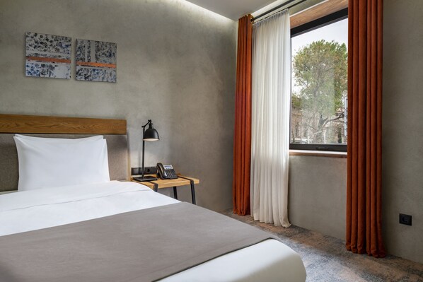 A hotel room with a large bed, a bedside table with a lamp, and a window with orange curtains.