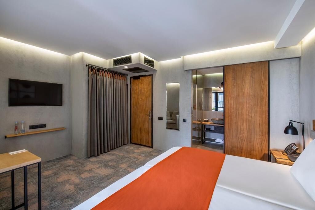 A modern hotel room with a bed, a desk, a TV mounted on the wall, and a bathroom visible through an open door.