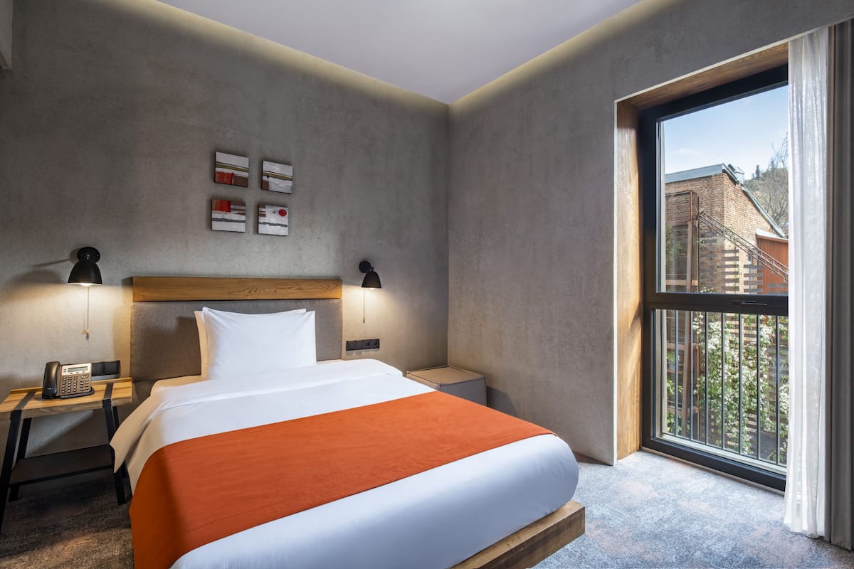 A hotel room with a large bed, a wooden headboard, a nightstand with a telephone, and a window with a view of buildings.