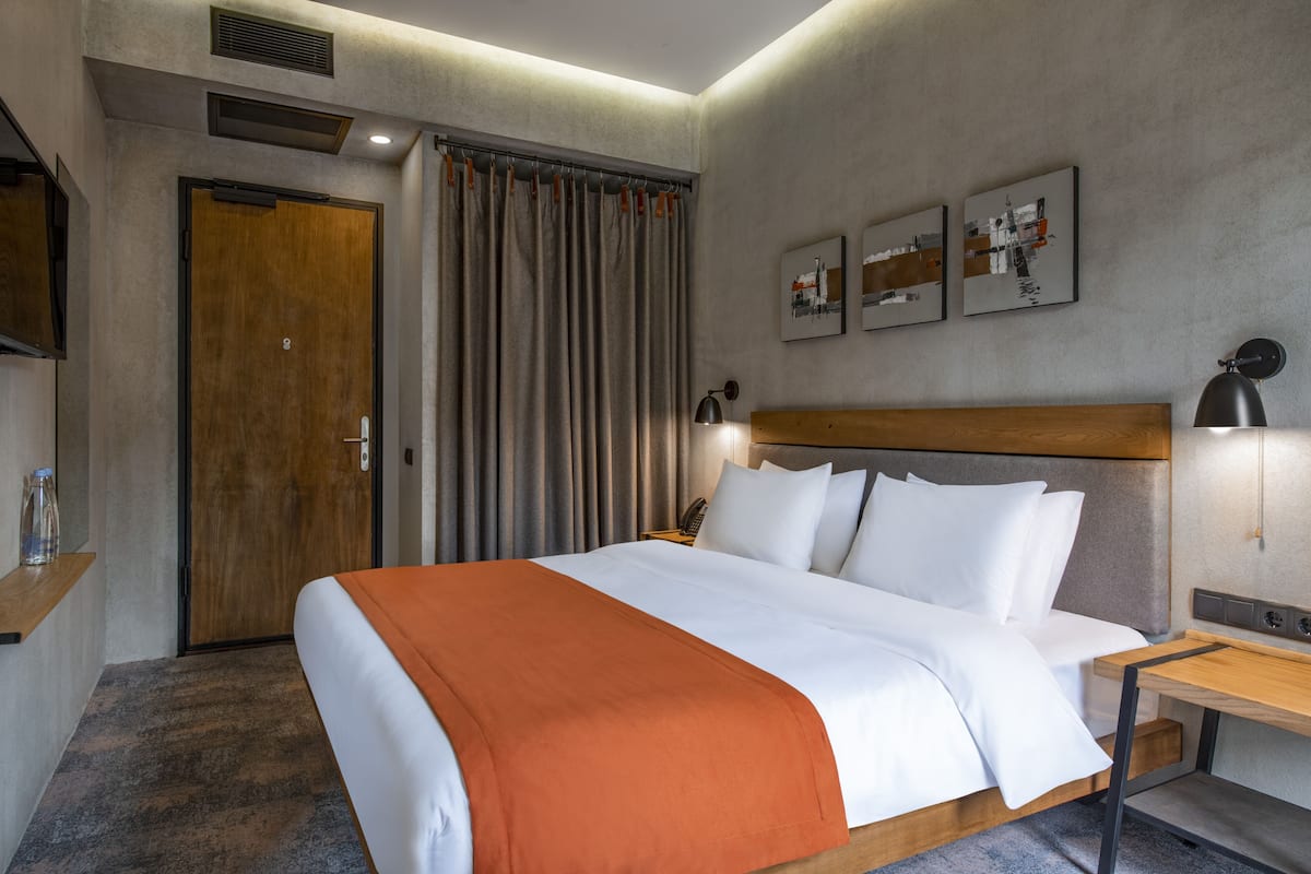 A hotel room with a bed, a wooden headboard, a nightstand, a desk, a lamp, a curtain, a door, and artwork on the wall.