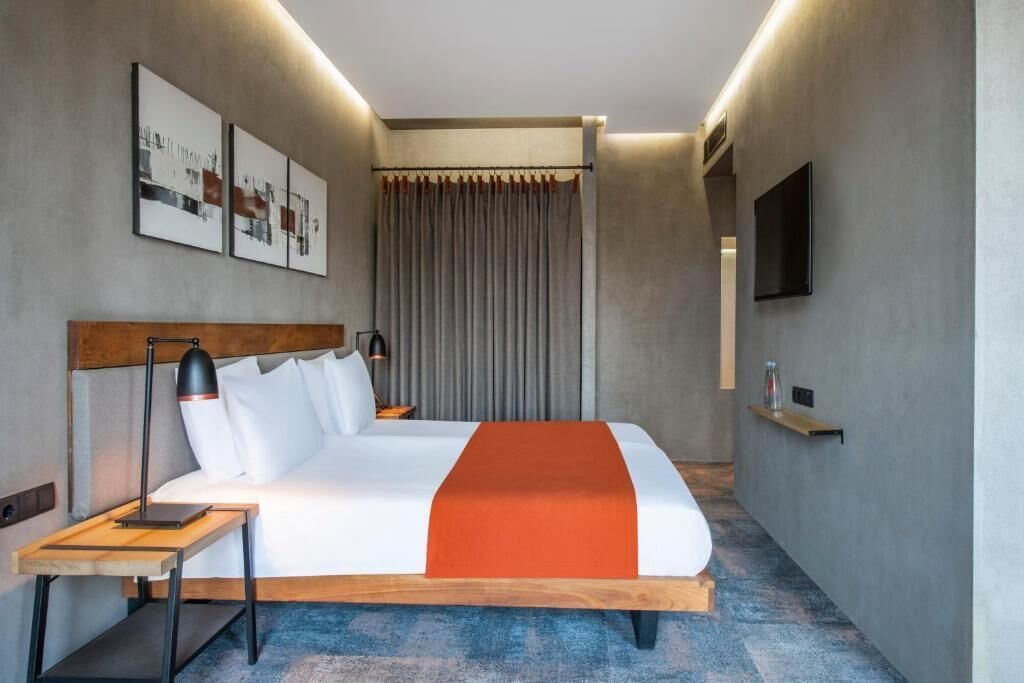 A modern hotel room with a large bed, a bedside table, a lamp, a TV, and a curtain.