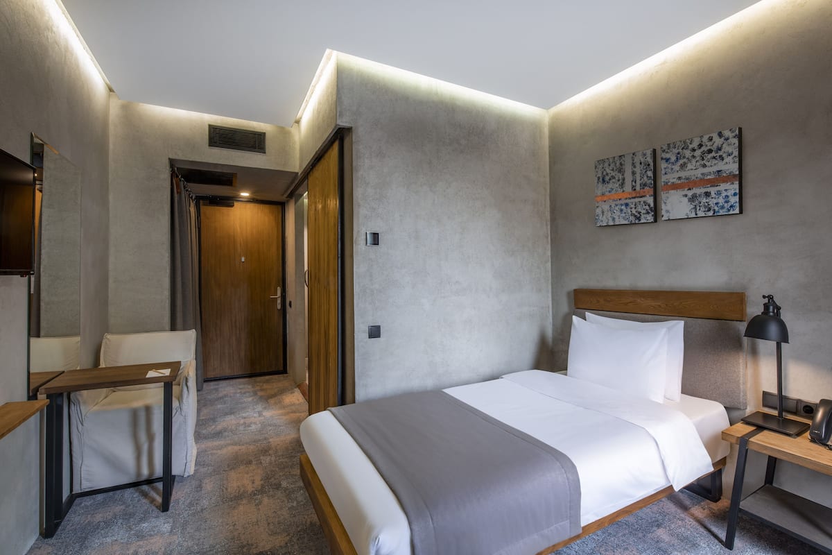 A hotel room with a bed, a desk, a chair, a nightstand, and a door.