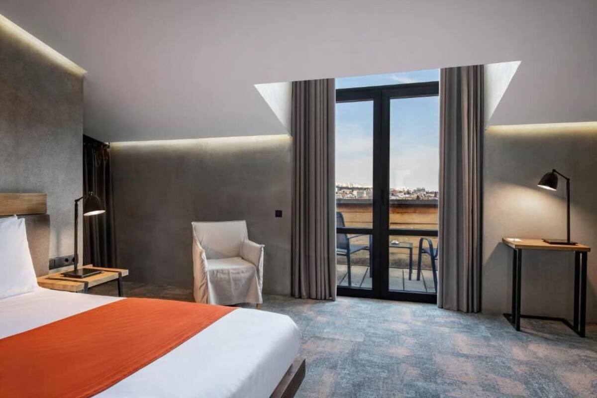 A hotel room with a bed, a chair, a desk, and a view of the city through a large window.