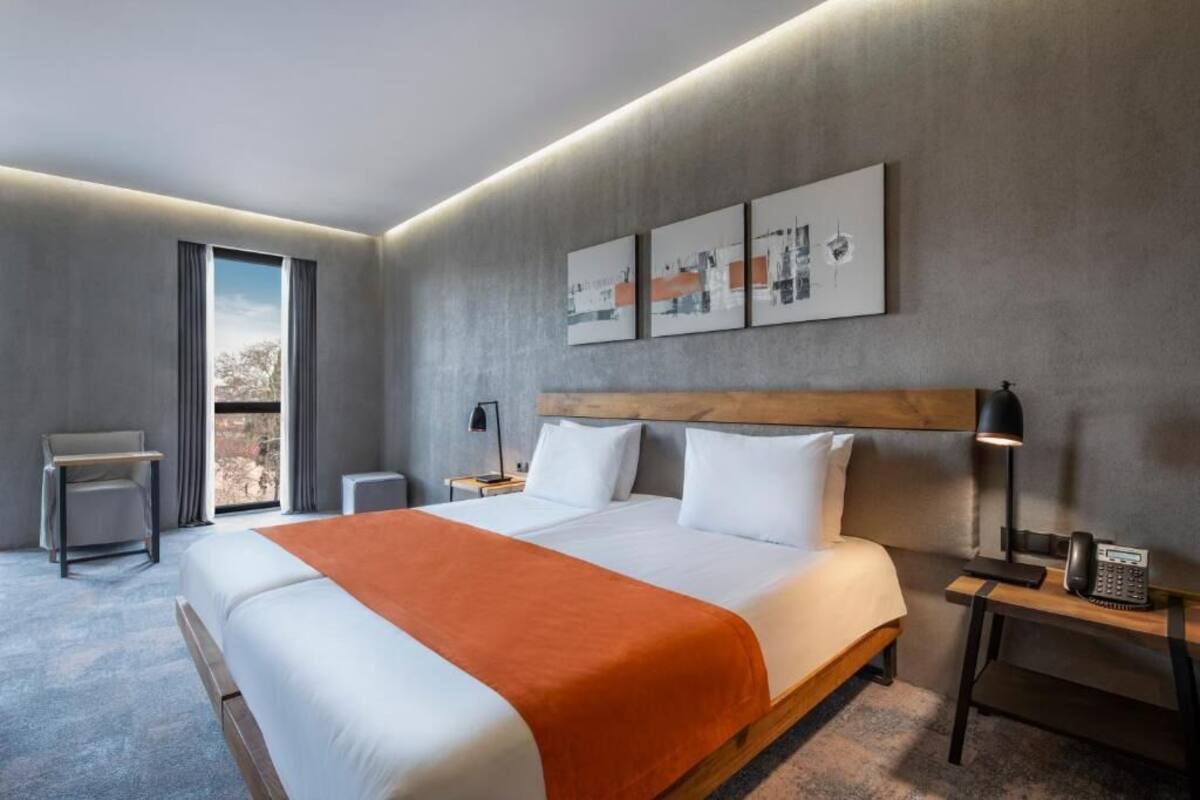 A modern hotel room with a large bed, a desk, a chair, and a view of the outdoors.