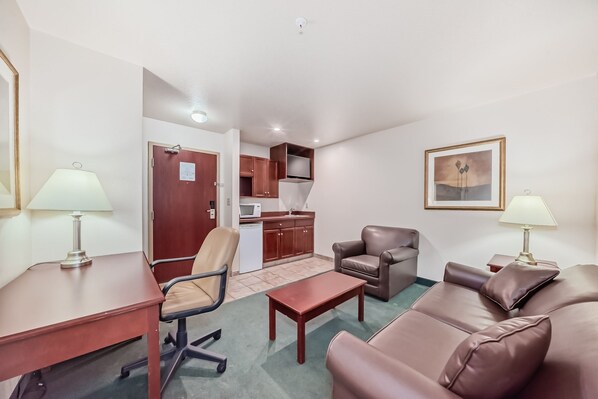 A compact living space with a kitchenette, a brown leather sofa, a brown leather armchair, a wooden coffee table, and a desk with a lamp. - Grande Cache Inn and Suites (Grande Cache)