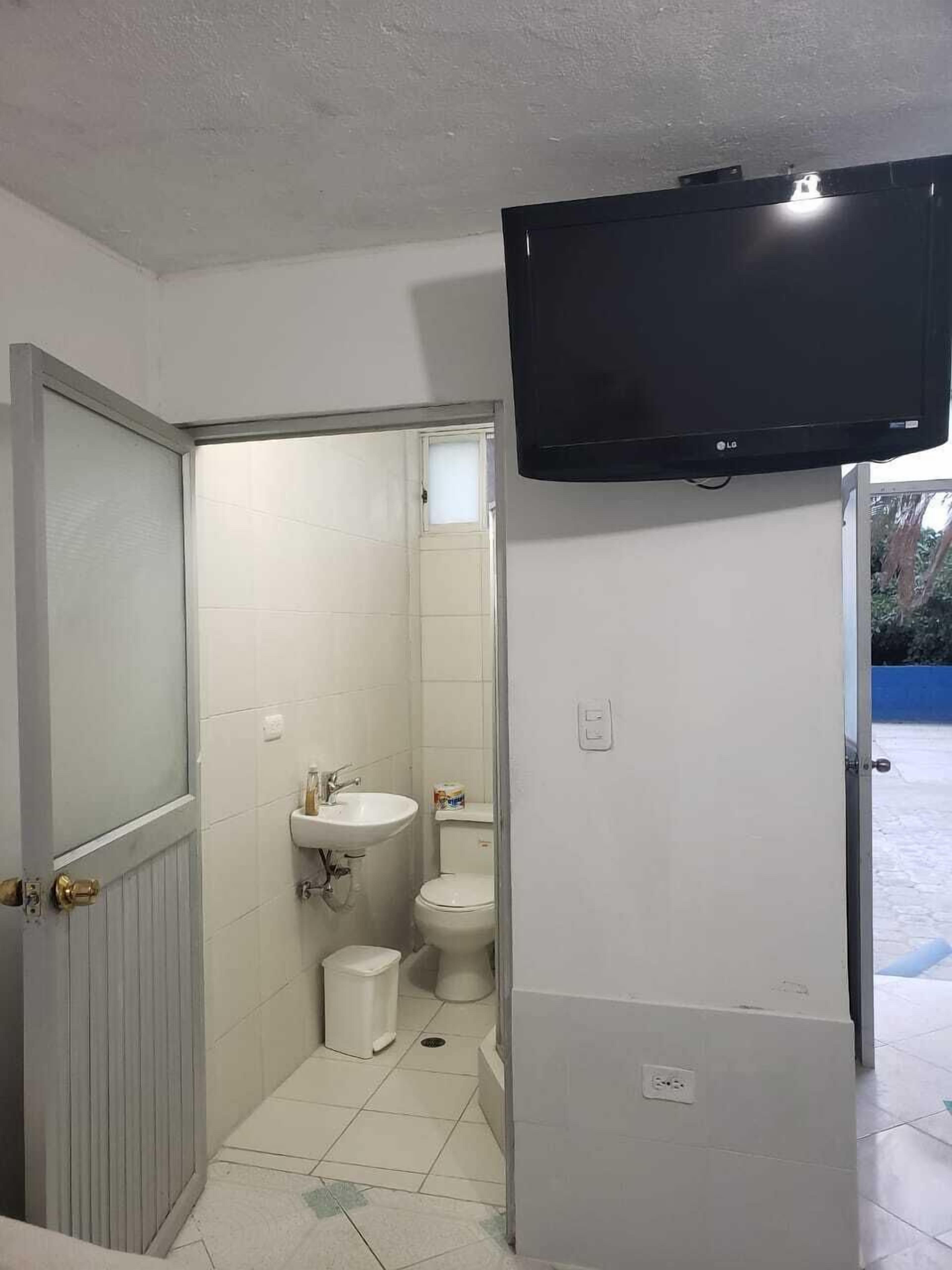 Une petite salle de bain comprenant des toilettes, un lavabo et une petite poubelle. Un téléviseur est fixé au mur, au-dessus du lavabo.
