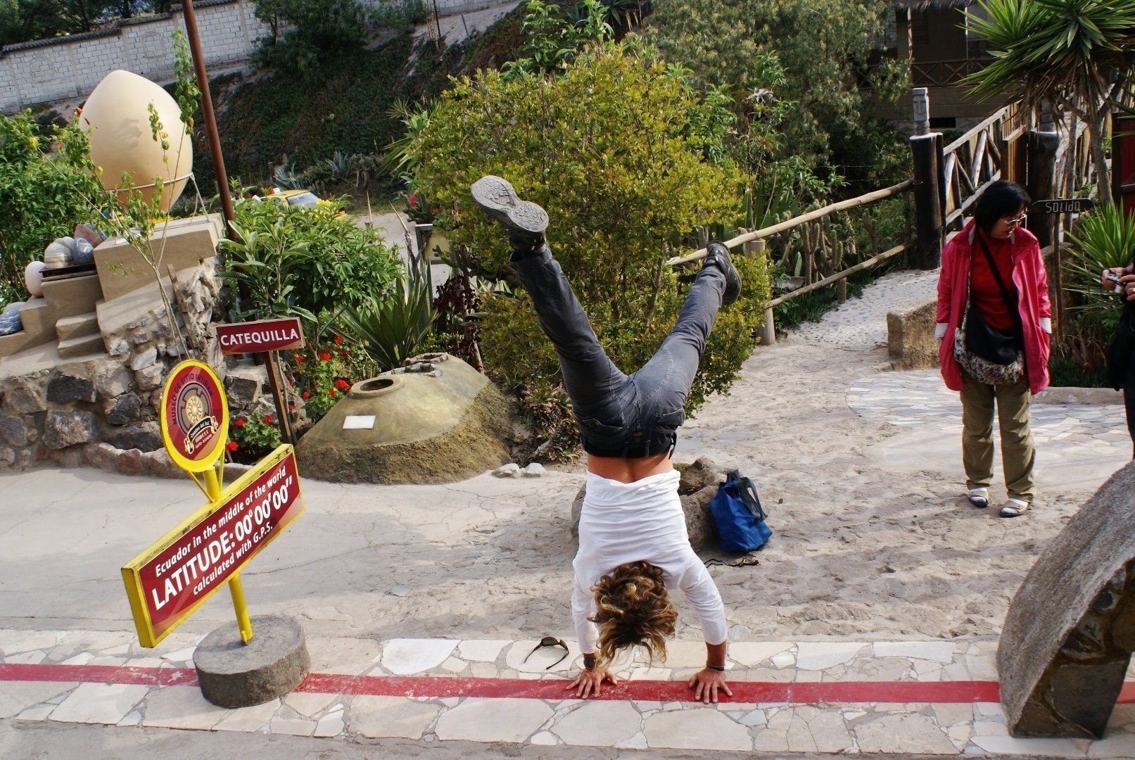 Una persona haciendo una postura de parada de manos sobre un sendero de piedra con un cartel que indica la latitud del lugar.